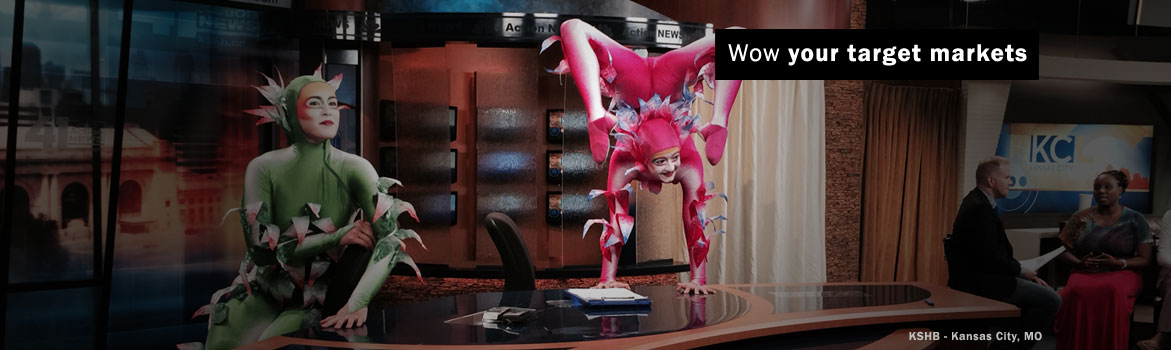 Two performers in colorful, elaborate costumes entertain on a television news set, while seated guests observe, highlighting a promotional event.