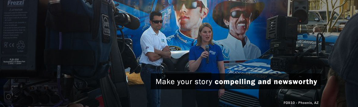 A woman speaks into a microphone at a NASCAR event, flanked by two men, with a promotional car and large images of famous racers in the background.