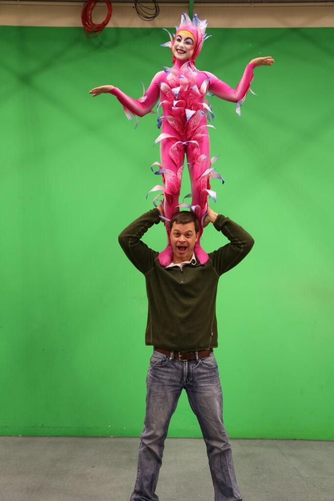 A performer dressed in a vibrant pink costume, adorned with colorful leaves, strikes a playful pose atop a man's shoulders against a green screen backdrop.