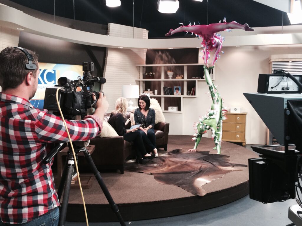 A television studio scene shows two women seated on a couch, discussing a topic, while a performer in a colorful plant-themed costume poses upside down in the background.