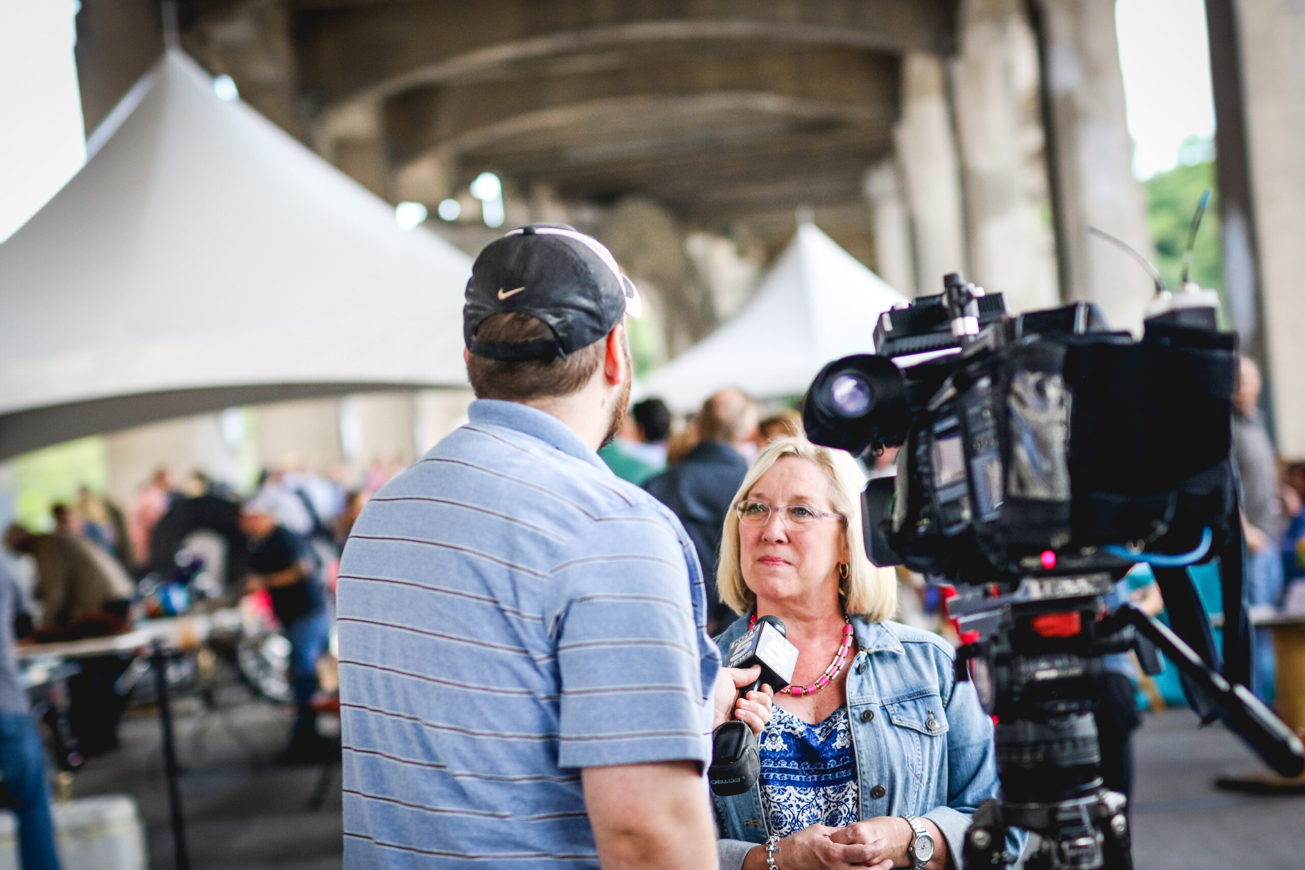 A woman with blonde hair speaks into a microphone during an interview under a large tent, with a camera focused on her.