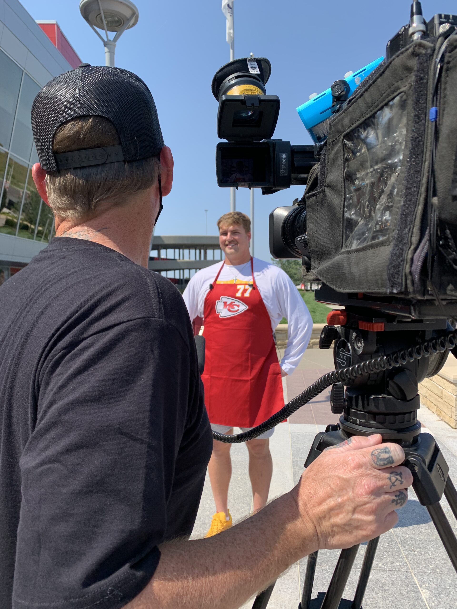 A sportsman in a red apron smiles while being interviewed outdoors, with a camera operator visible in the foreground, capturing the moment.