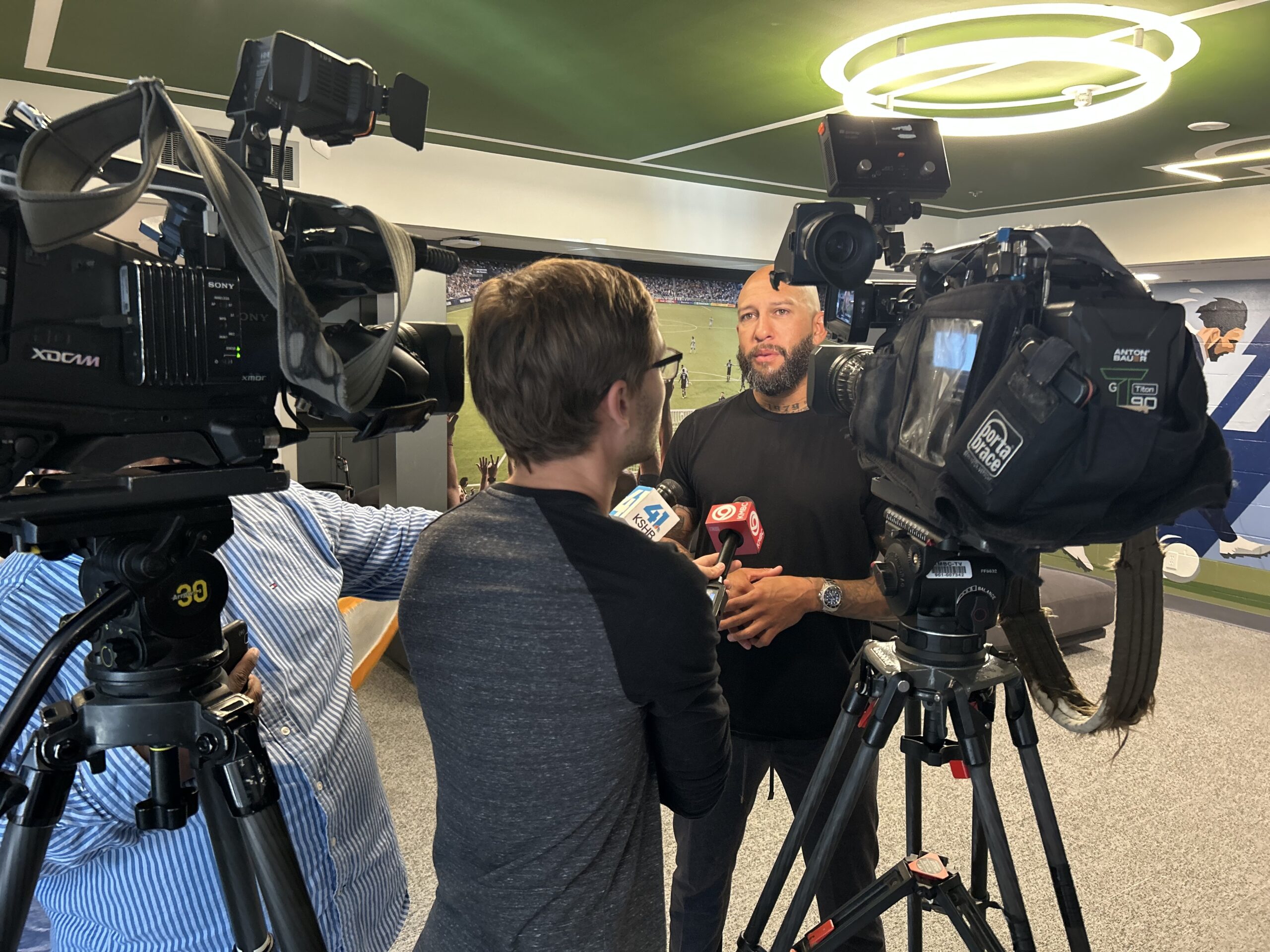 A man being interviewed by reporters in a sports setting, surrounded by cameras and a backdrop of a soccer field.