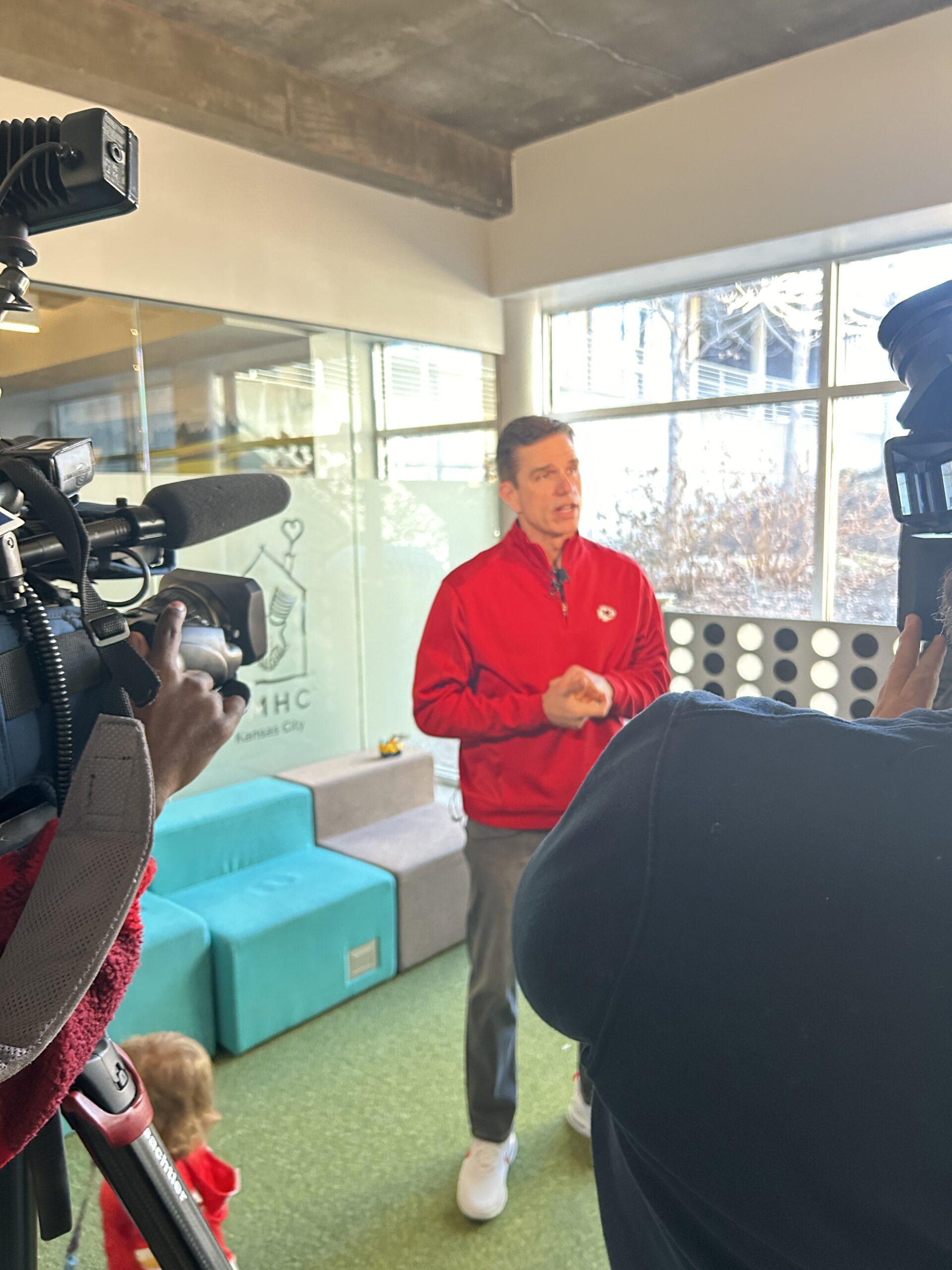 A man in a red jacket speaks to the camera in a brightly lit room. Children play in the background, emphasizing a community event or engagement.
