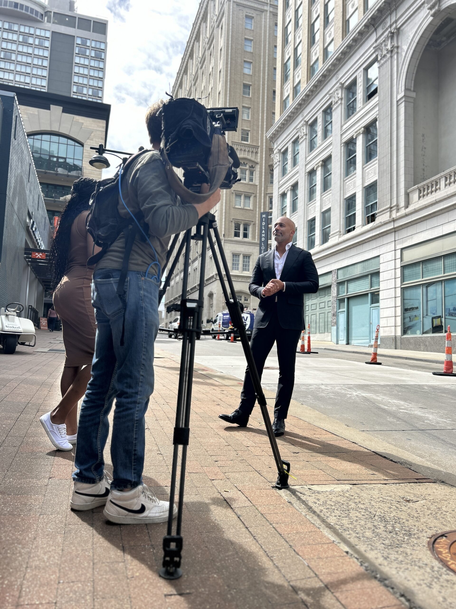 A news crew films a man in a suit speaking on a city street, while a woman stands nearby. The scene captures a professional interview setting.