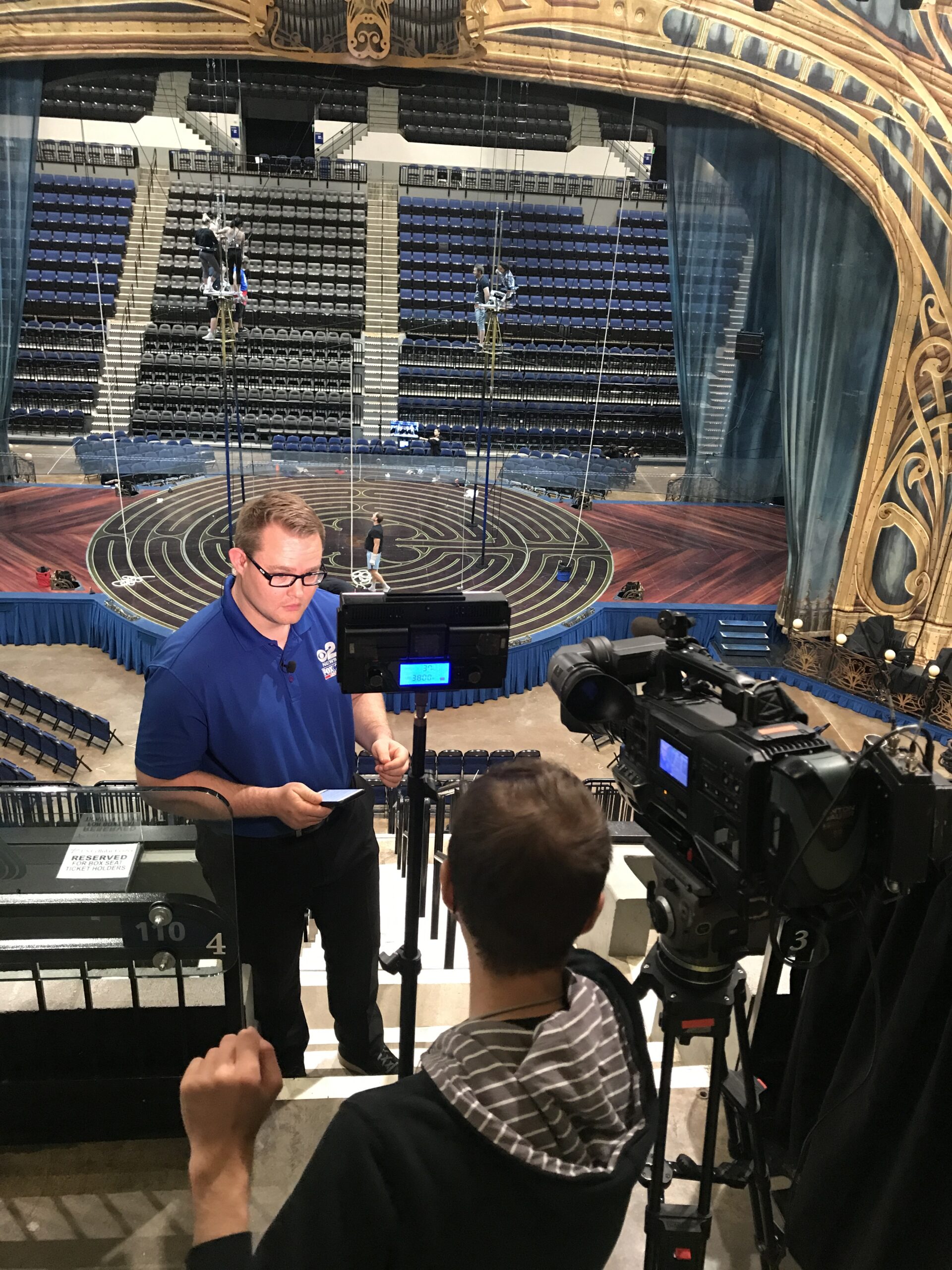 A presenter in a blue shirt holds notes while speaking to a camera crew in front of a partially set stage, with safety equipment and seating visible.