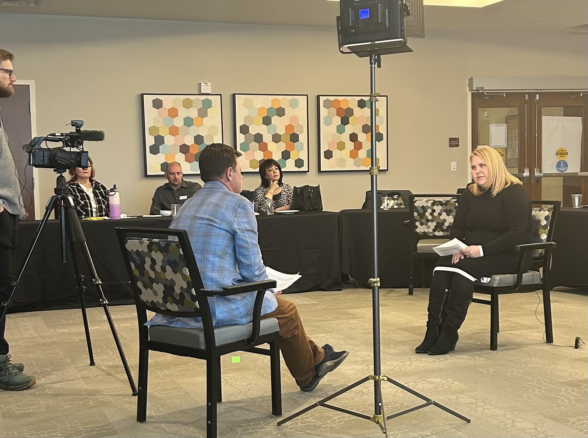 A formal interview scene featuring a woman in a black dress speaking to a man in a checked shirt, with a camera crew and audience present.
